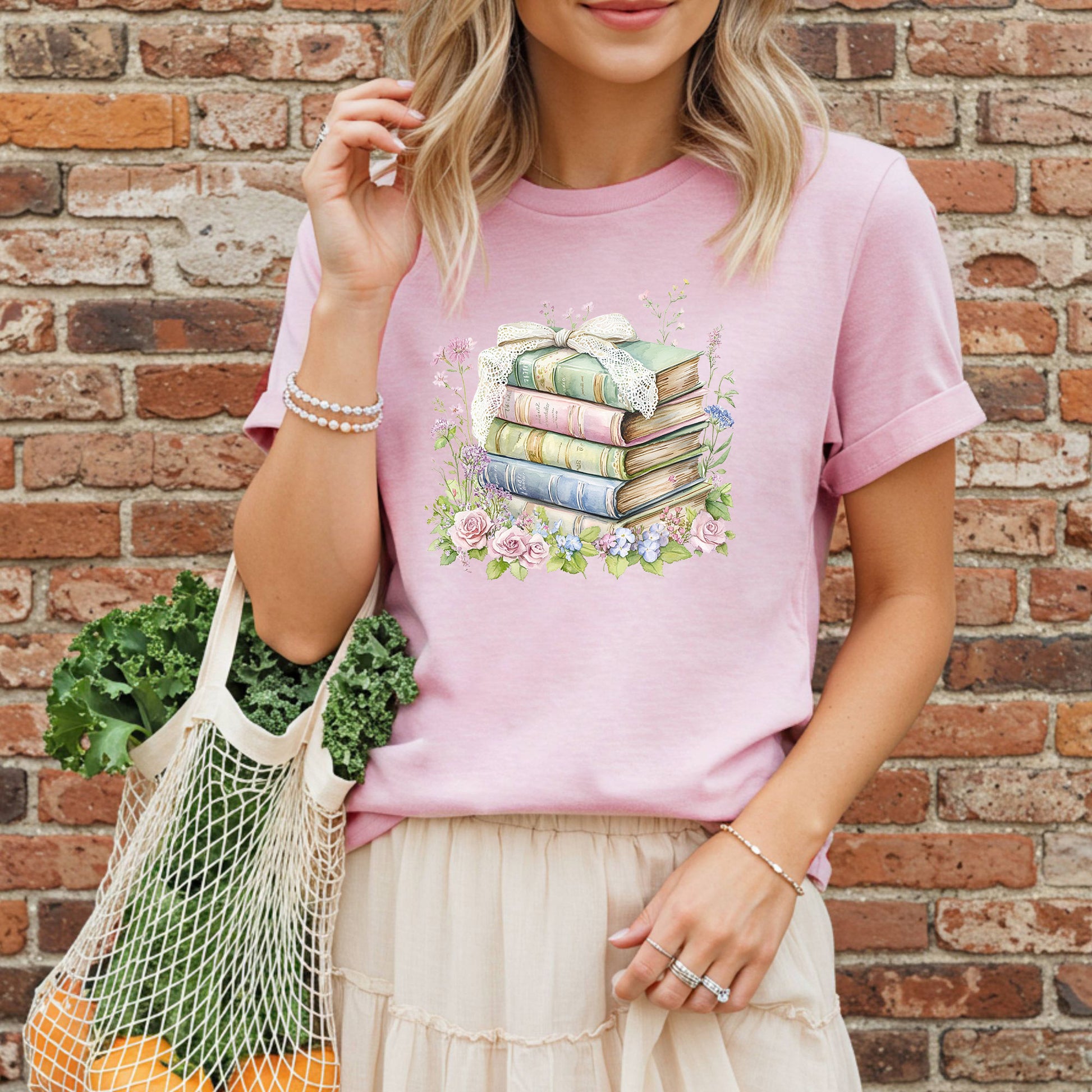 Pink t-shirt with a watercolor stack of vintage books, lace ribbon, and botanical flowers like roses and hydrangeas.