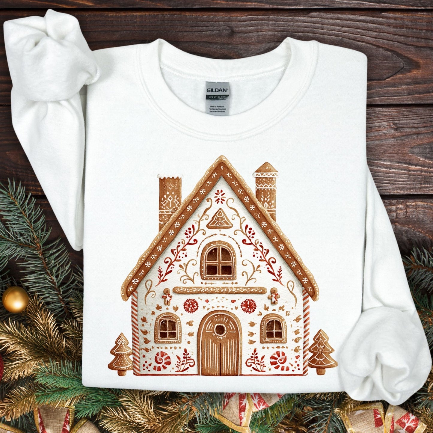 White Gildan sweatshirt with a folk art gingerbread house design, surrounded by festive greenery.