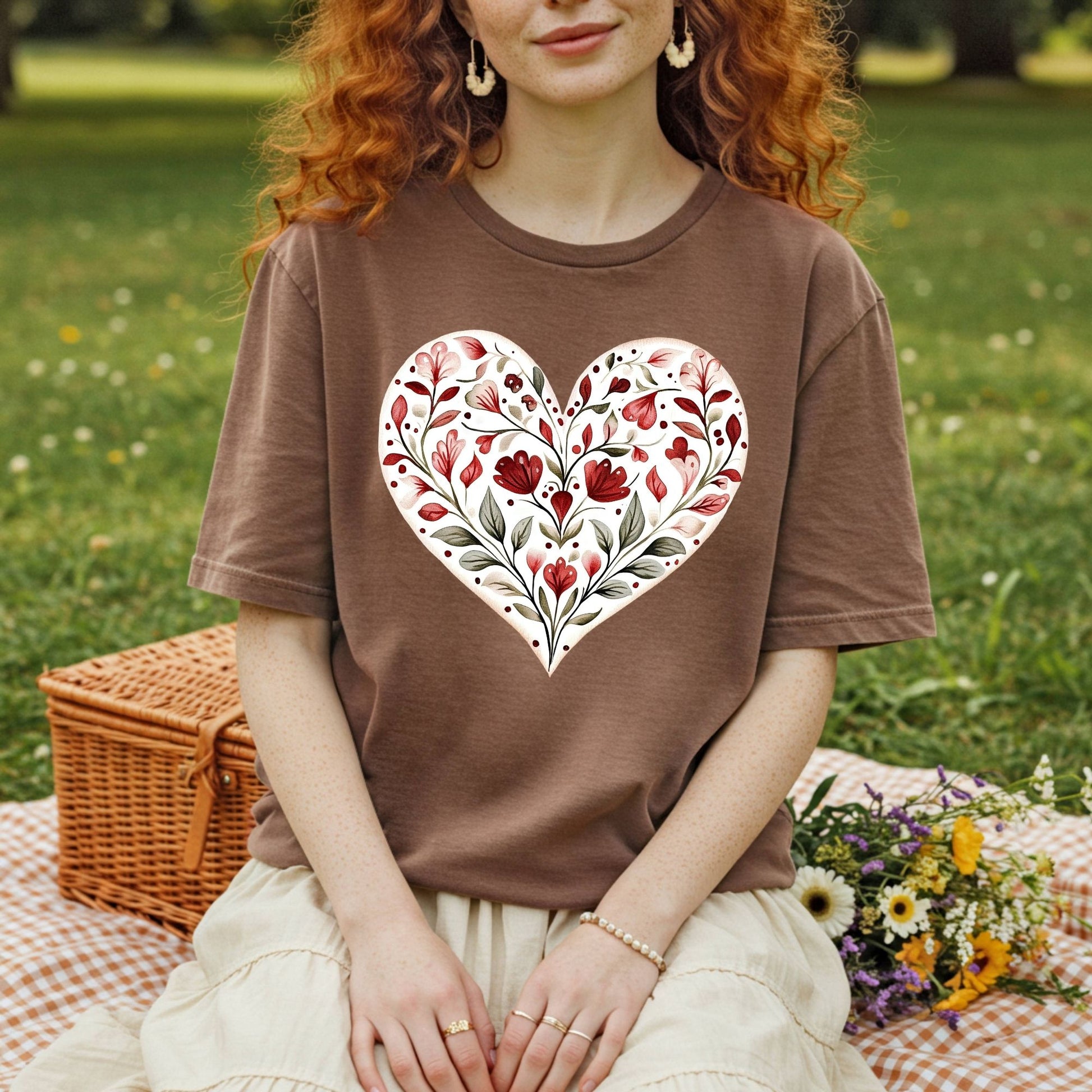 Brown relaxed-fit t-shirt with a red and green folk-inspired floral heart design, worn by a person in a park.