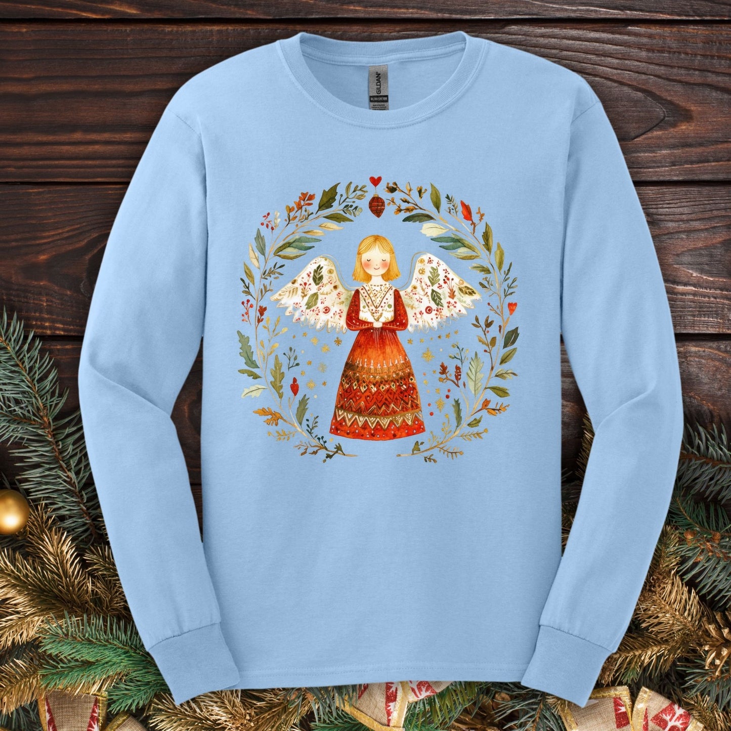 Light blue long sleeve tee with a folk art angel in a winter wreath, featuring red, green, and gold accents.