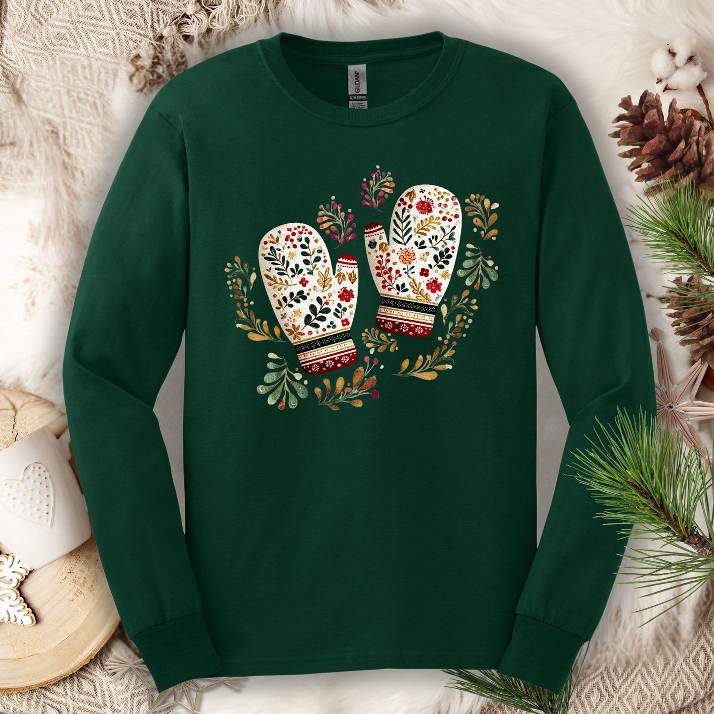 Forest Green long sleeve tee with folk art watercolor mittens in red, cream, evergreen, and gold, surrounded by winter foliage.