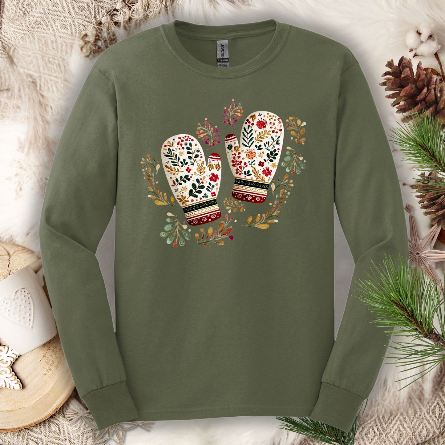 Military green long sleeve tee with watercolor folk art mittens in red, cream, evergreen, and gold, surrounded by winter foliage.