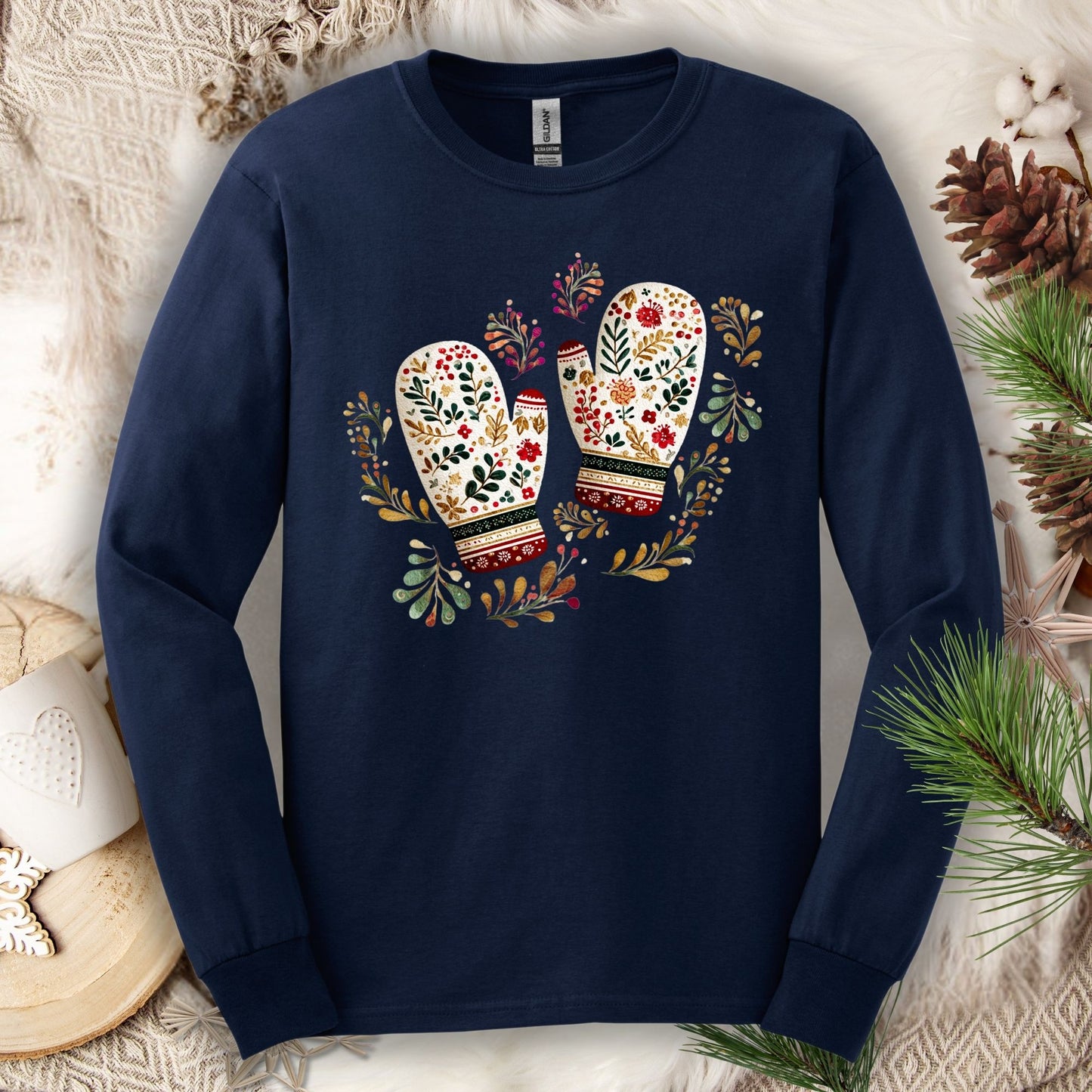 Navy long sleeve tee with watercolor folk art mittens in red, cream, evergreen, and gold, surrounded by winter foliage.