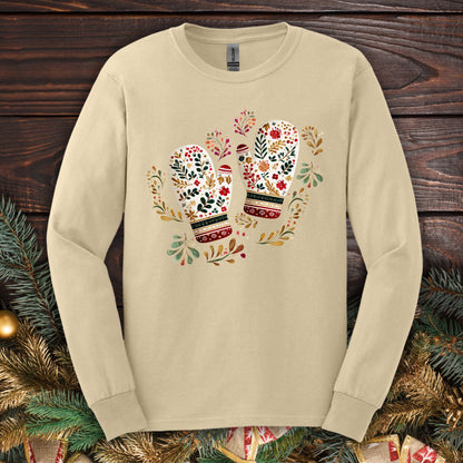 Sand long sleeve tee with watercolor folk art mittens in red, cream, evergreen, and gold, surrounded by winter foliage.