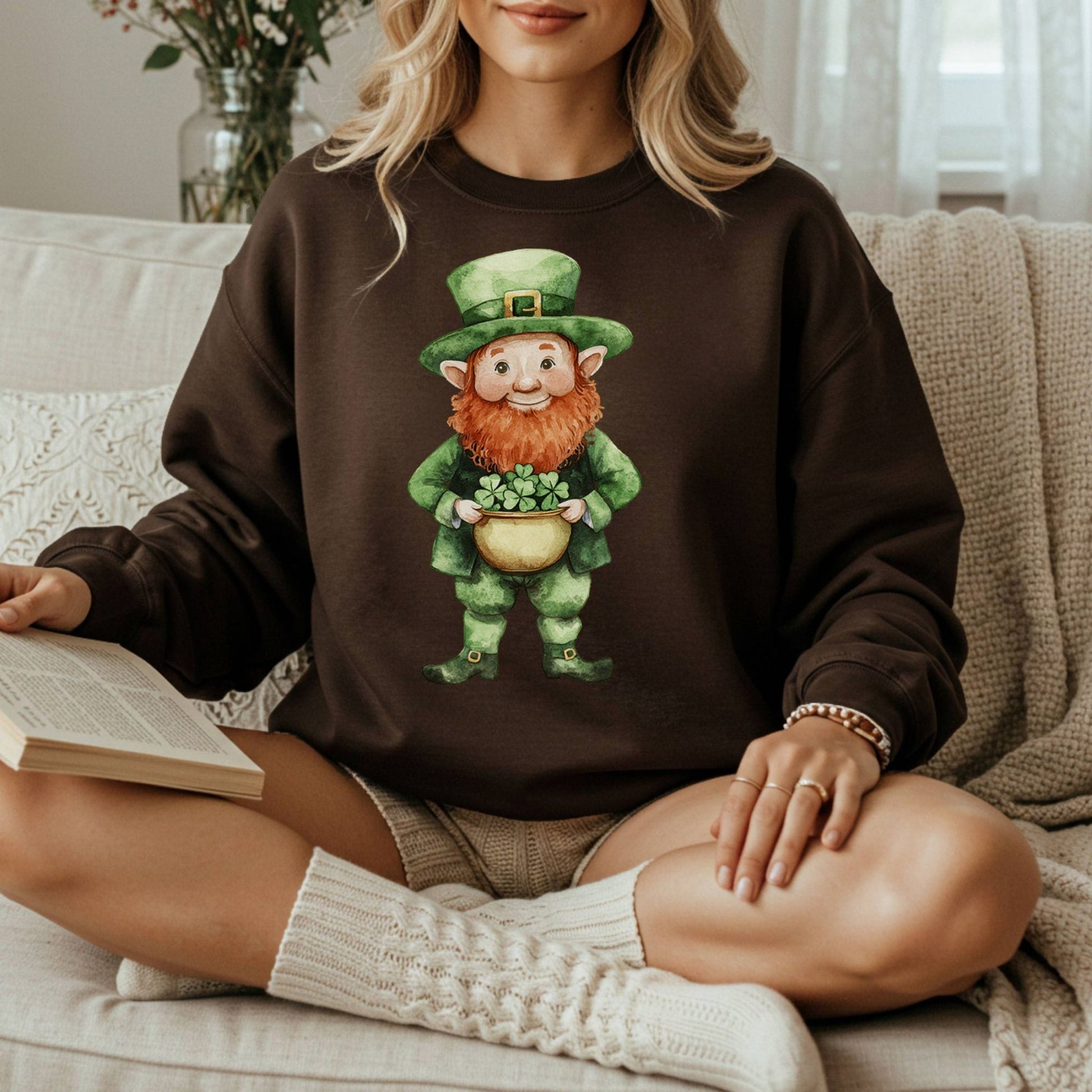 Unisex brown sweatshirt with a watercolor leprechaun holding a four-leaf clover, worn by a woman reading.