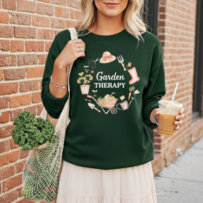 Woman in a dark green "Garden Therapy" sweatshirt with a floral wreath design, holding a coffee and mesh bag of greens.