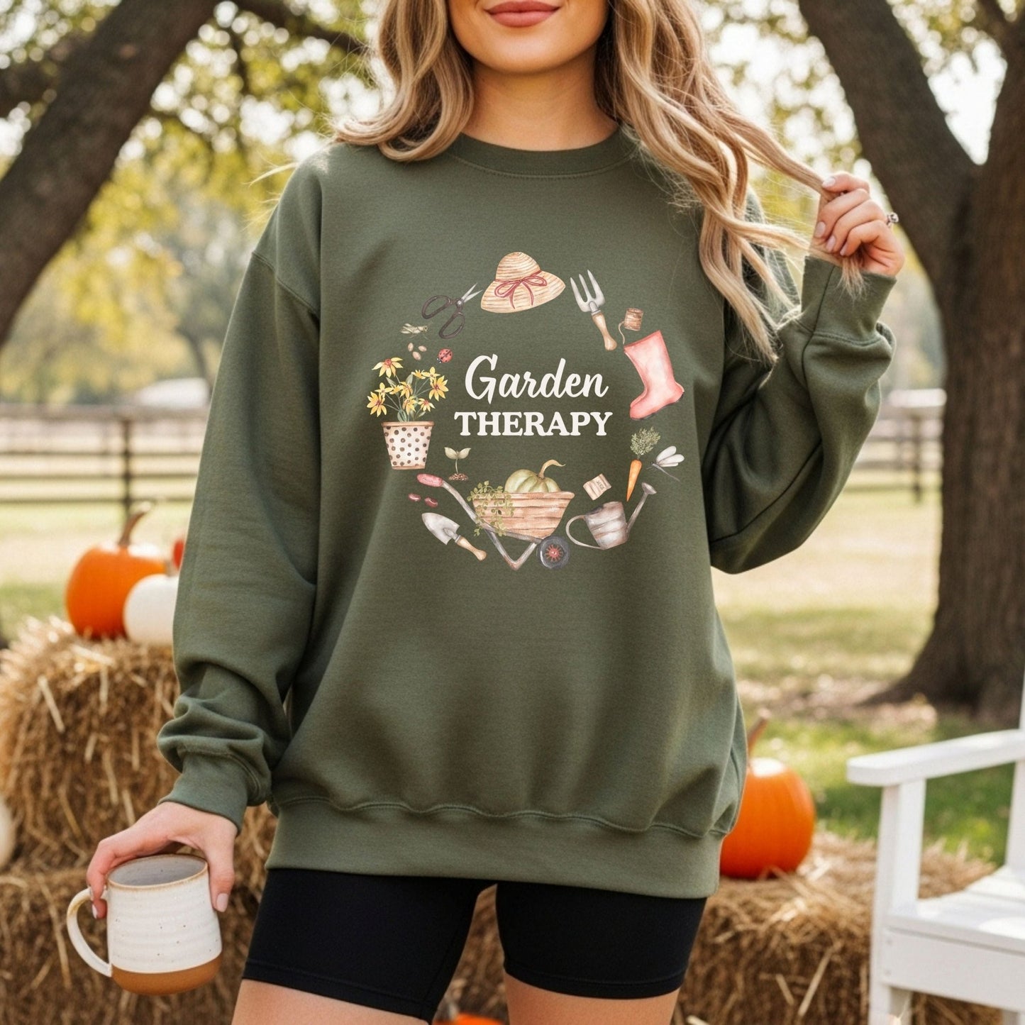 Woman in a green "Garden Therapy" sweatshirt with a floral wreath of gardening tools, holding a mug.