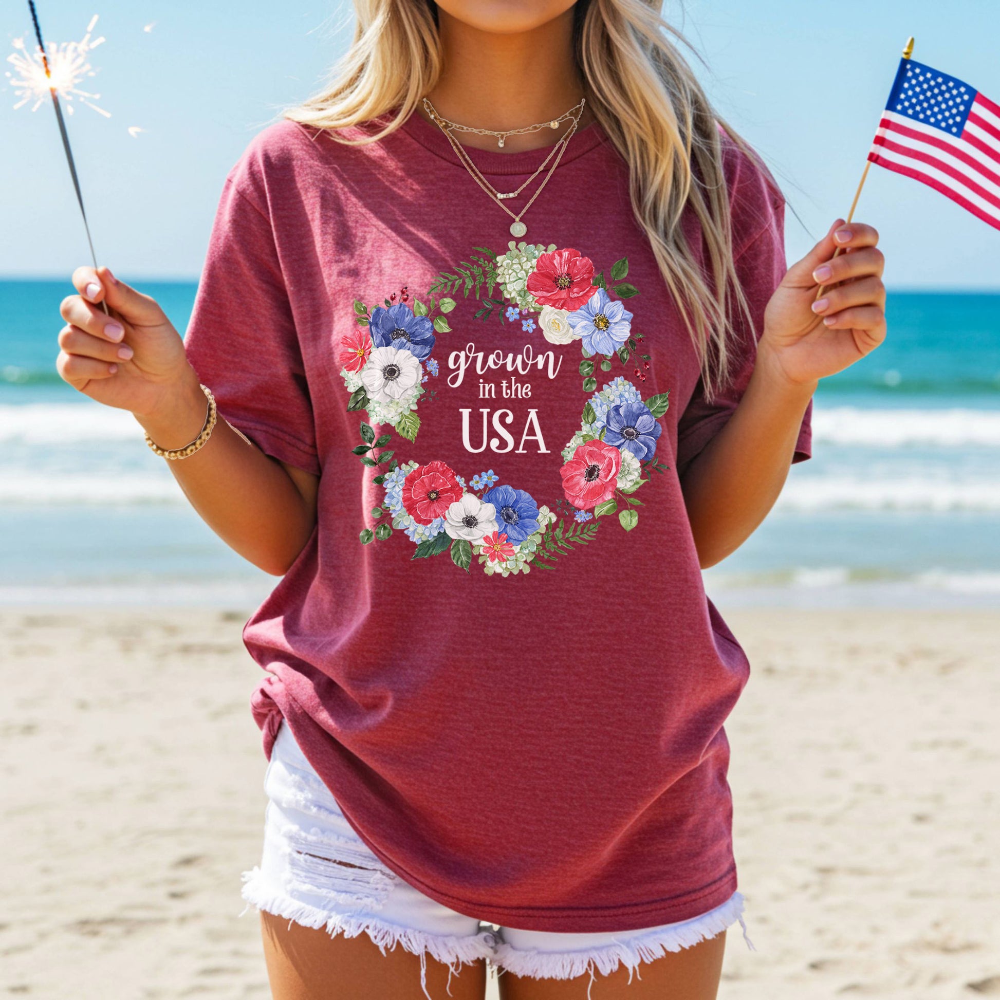 Red t-shirt with a watercolor floral wreath and "Grown in the USA" text, held by a person on a beach.