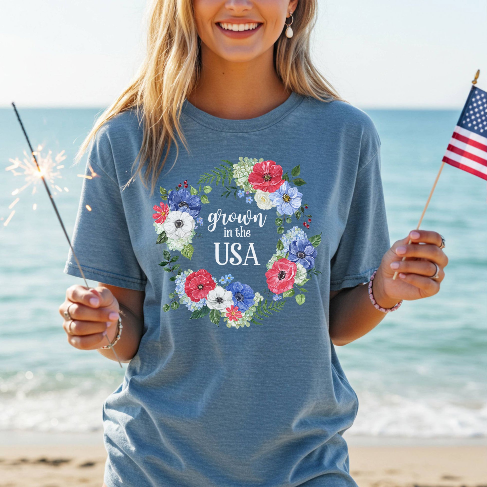 Woman in a blue "Grown in the USA" t-shirt with a floral wreath, holding a sparkler and American flag.