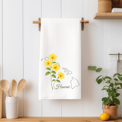 White microfiber tea towel with yellow hibiscus flowers and a subtle Hawaii map, hanging on a wooden rack.