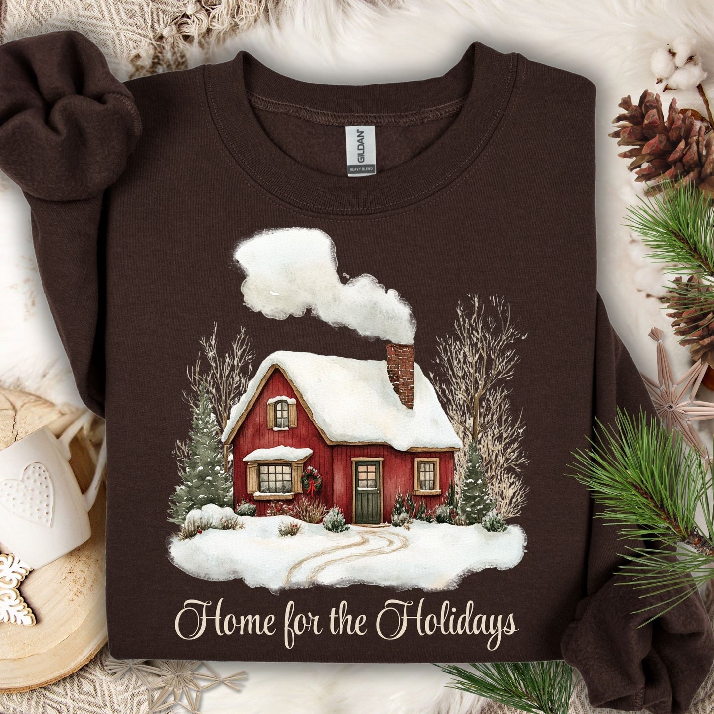 Dark chocolate Gildan sweatshirt with vintage watercolor red cabin, snow, evergreens, and "Home for the Holidays" text.