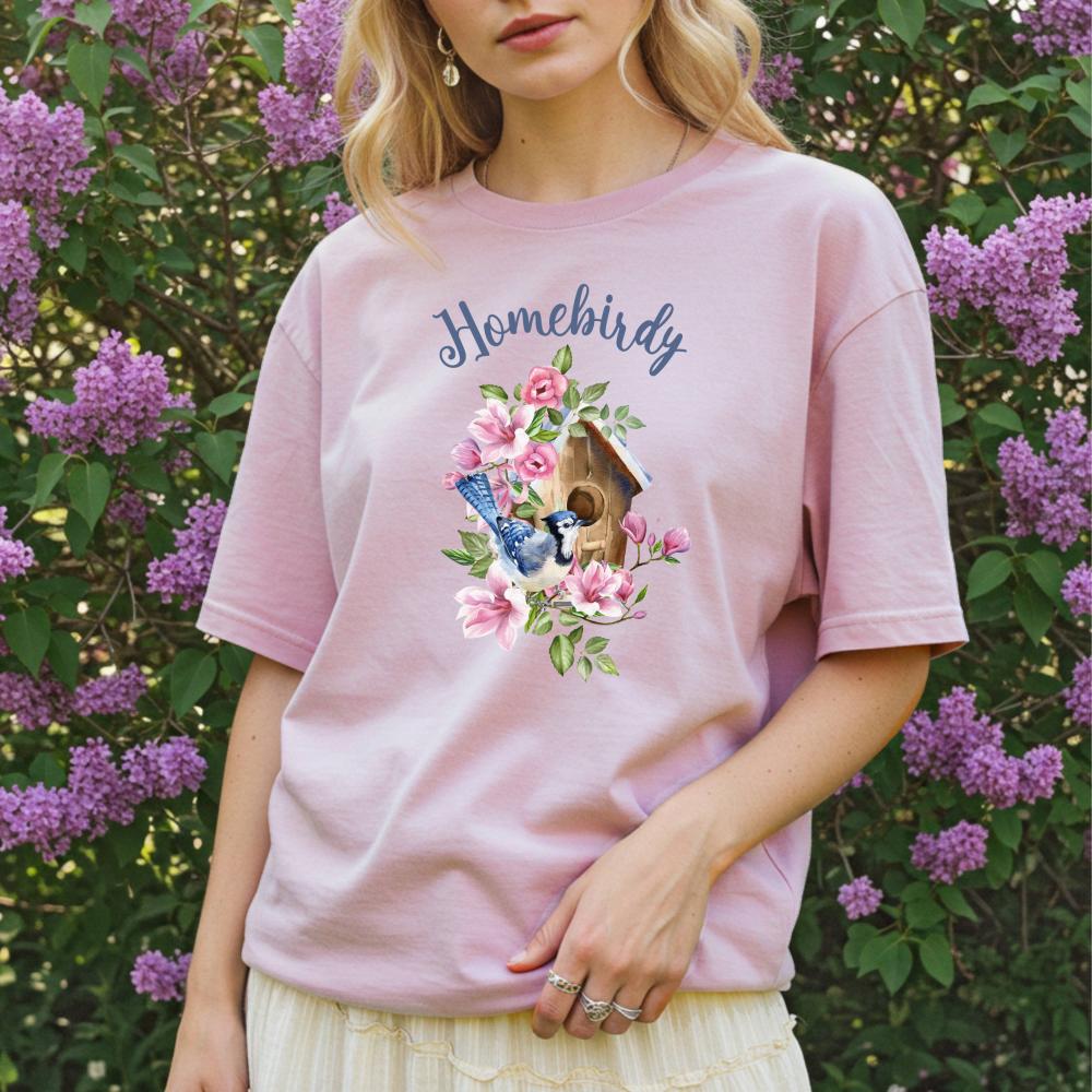 Person wearing a pink 'Homebirdy' t-shirt with a birdhouse design in front of purple flowers.