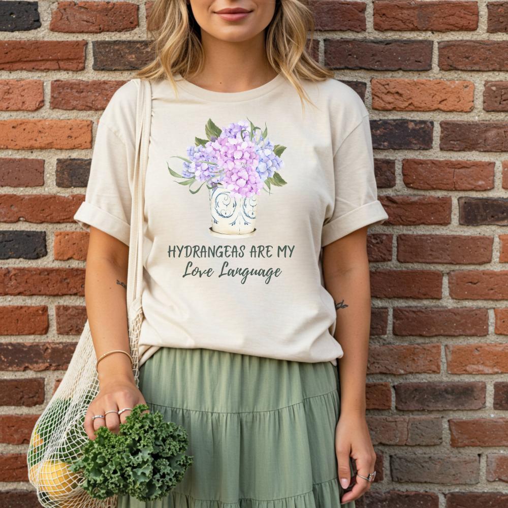 woman wearing a t-shirt with a floral design with text that reads "Hydrangeas are my love language" standing against a brick wall.