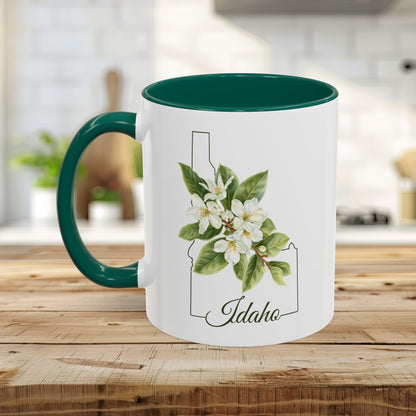 White ceramic mug with green handle and interior, featuring Idaho state silhouette and Syringa flowers.