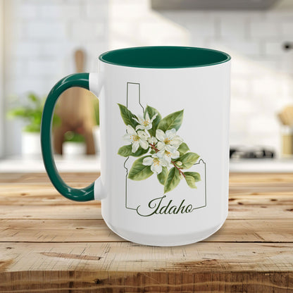 White ceramic mug with green handle/interior, Idaho state outline, and Syringa floral illustration.