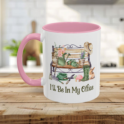 White ceramic 11oz mug with pink handle and interior, featuring watercolor gardening tools, bench, and "I'll Be In My Office" text.