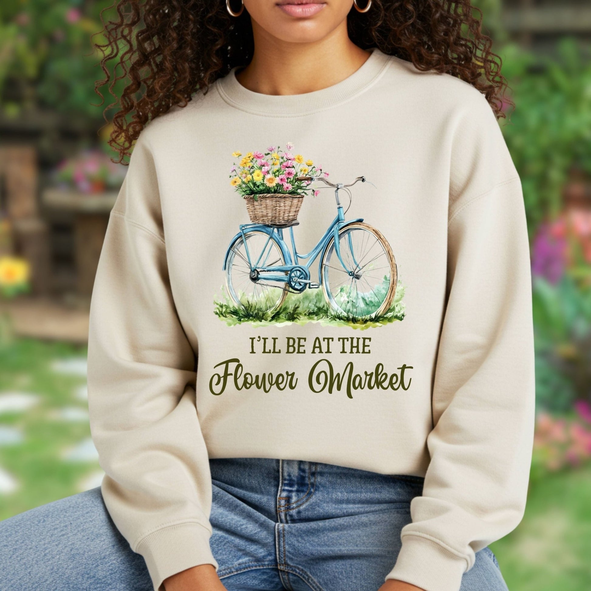 Beige crewneck sweatshirt with a blue bicycle, flower basket, and "I'll be at the Flower Market" text.