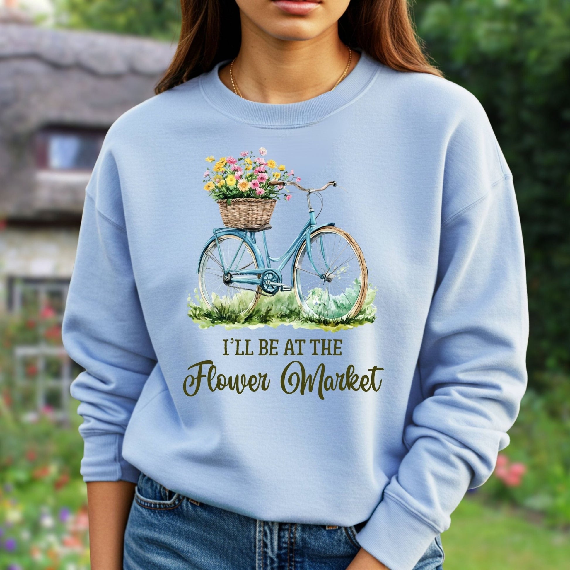 Light blue crewneck sweatshirt with a hand-illustrated bicycle, flower basket, and "I'll be at the Flower Market" text.
