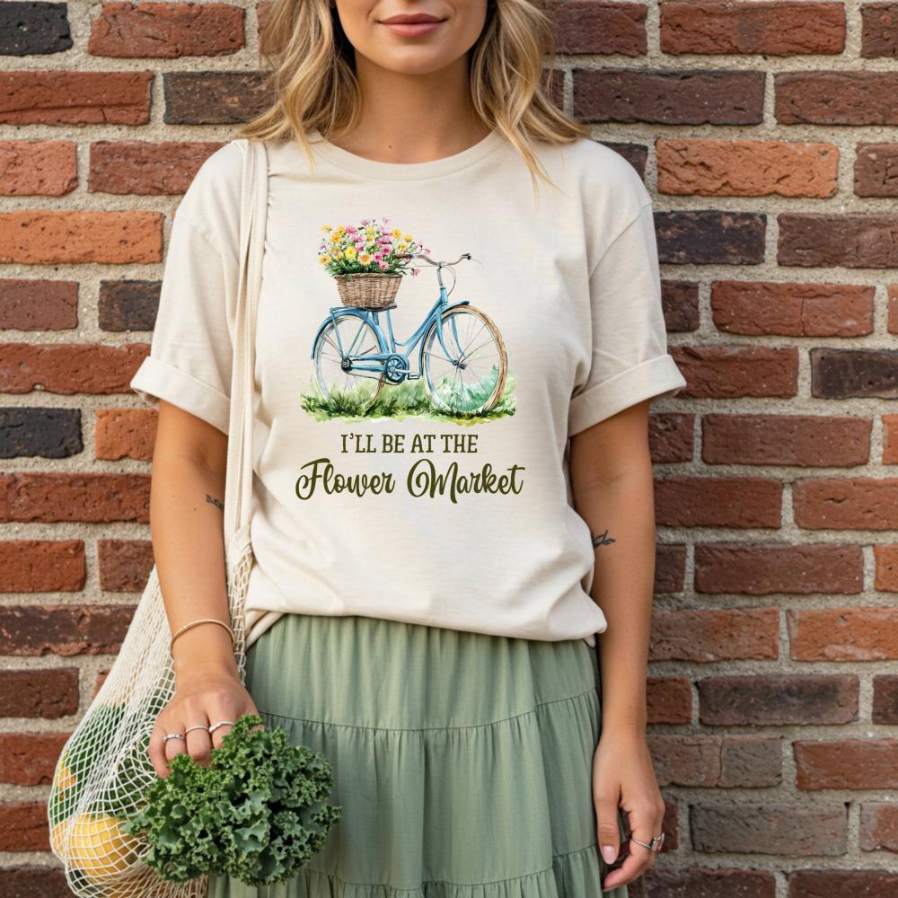 woman wearing a t-shirt with a bicycle and flower market graphic that reads "I'll be at the flower market", standing against a brick wall.