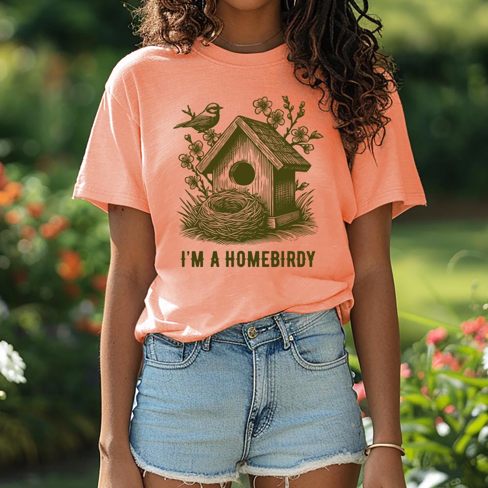 young woman wearing a peach t-shirt with a birdhouse graphic and text, standing outdoors.