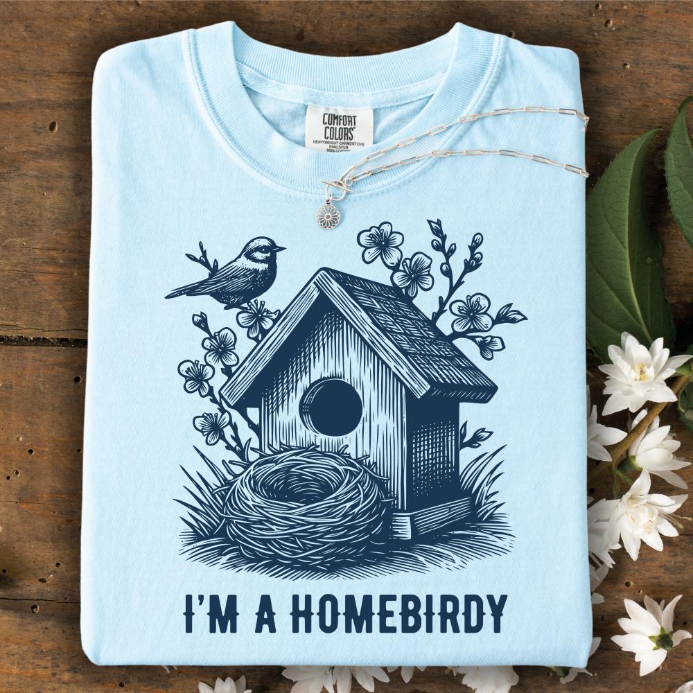 Light blue t-shirt with birdhouse and nest design, text 'I'm a Homebirdy', on a wooden surface with flowers.