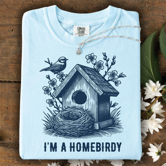 Light blue t-shirt with birdhouse and nest design, text 'I'm a Homebirdy', on a wooden surface with flowers.