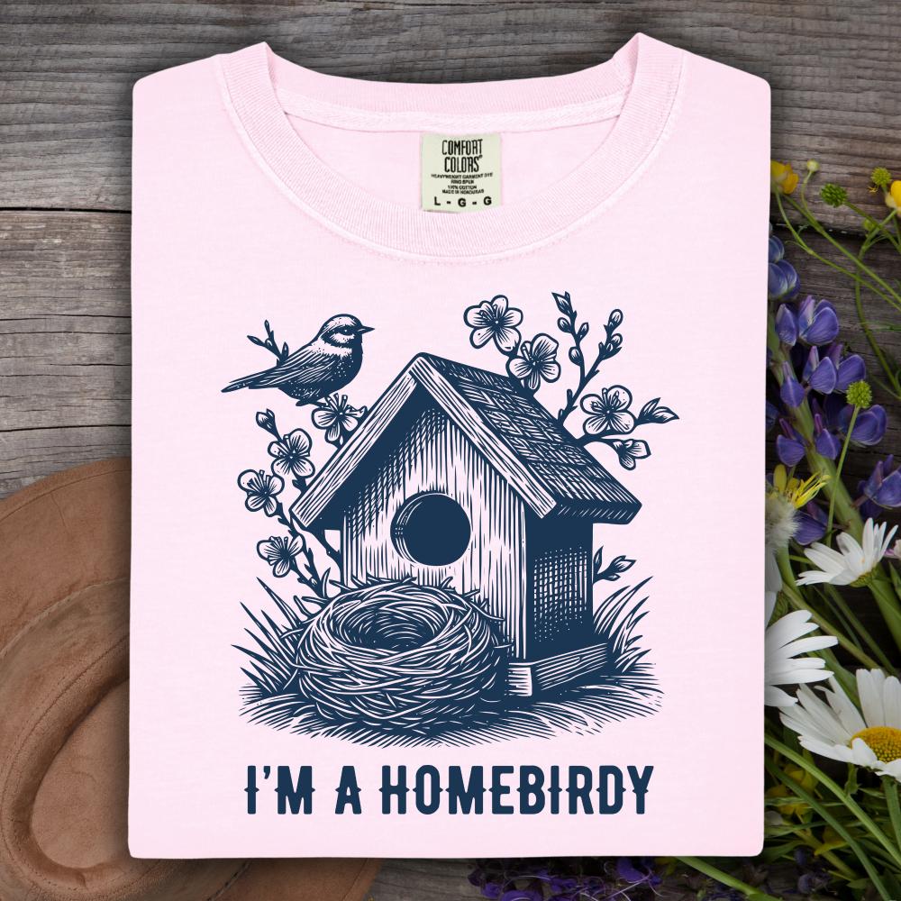 Pink t-shirt with a birdhouse and nest design and text 'I'm a Homebirdy' on a wooden surface with flowers.