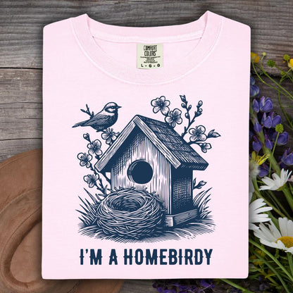 Pink t-shirt with a birdhouse and nest design and text 'I'm a Homebirdy' on a wooden surface with flowers.