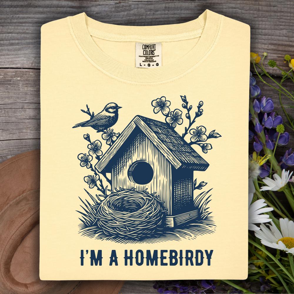 light yellow t-shirt with a birdhouse and nest design and 'I'm a Homebirdy' text, placed on a wooden surface with flowers.