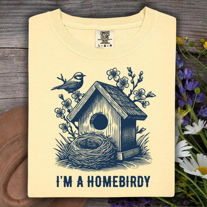 light yellow t-shirt with a birdhouse and nest design and 'I'm a Homebirdy' text, placed on a wooden surface with flowers.
