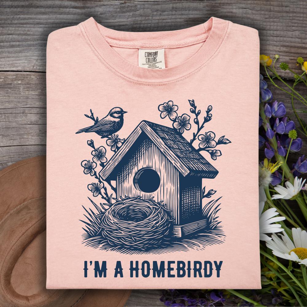 Pink t-shirt with a birdhouse and nest design and 'I'm a Homebirdy' text, placed on a wooden surface with flowers.