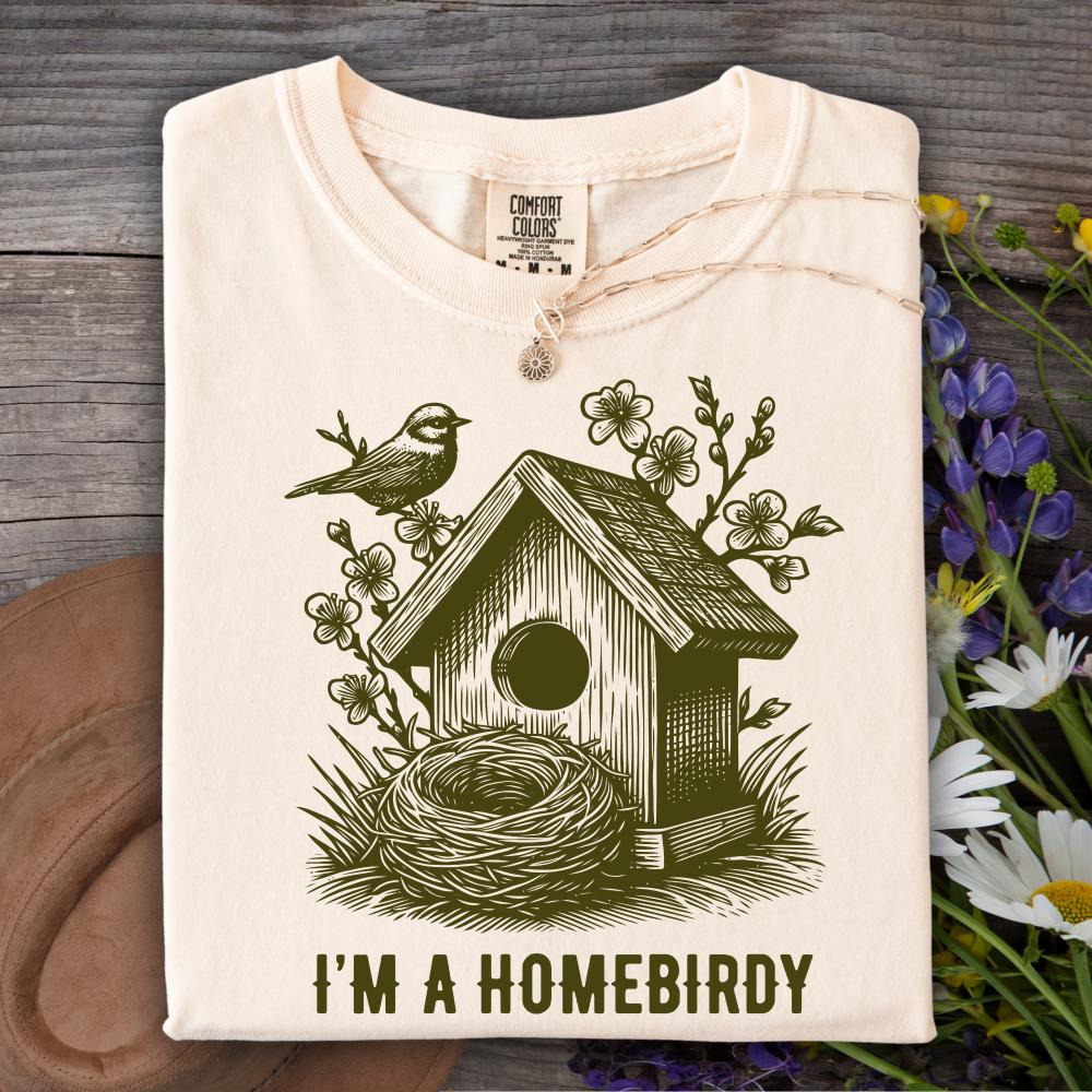 Folded ivory t-shirt with a birdhouse and nest design and 'I'm a Homebirdy' text, placed on a wooden surface with flowers.