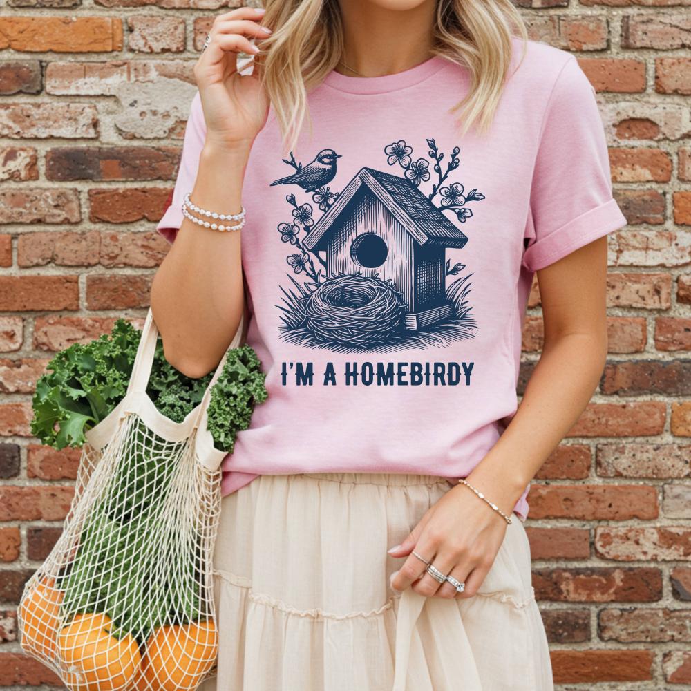young woman wearing a pink t-shirt with a birdhouse design and text, standing against a brick wall.