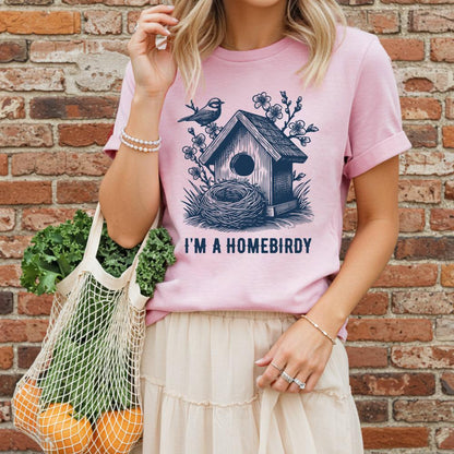 young woman wearing a pink t-shirt with a birdhouse design and text, standing against a brick wall.