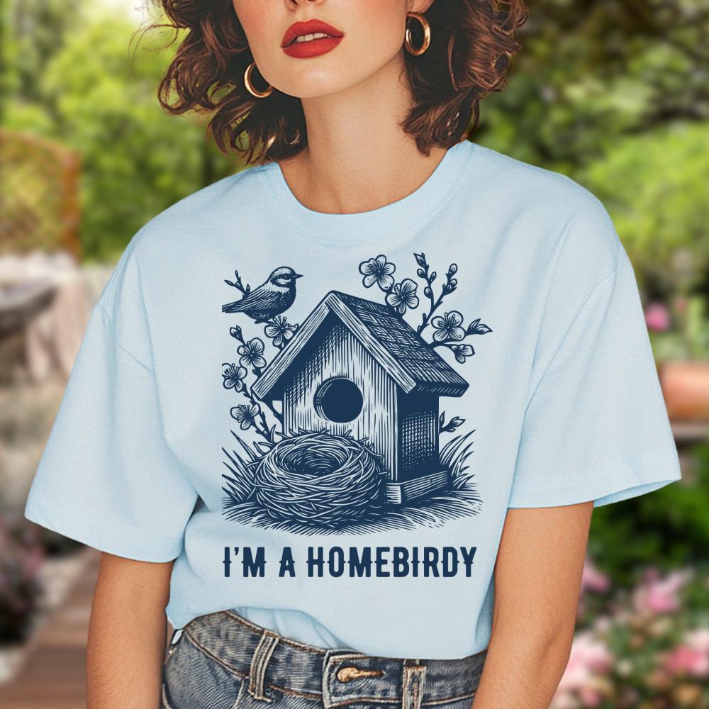 young woman wearing a light blue t-shirt with a birdhouse and nest design, text 'I'm a Homebirdy', outdoors.