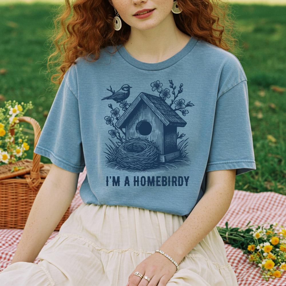 young woman with curly red hair wearing a blue t-shirt with a birdhouse design and text, sitting outdoors on a blanket.