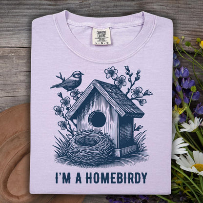 Light purple t-shirt with a birdhouse and nest design and 'I'm a Homebirdy' text, placed on a wooden surface with flowers.