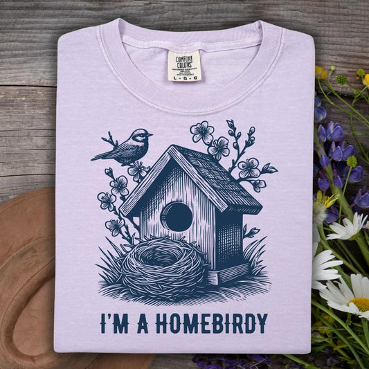 Light purple t-shirt with a birdhouse and nest design and 'I'm a Homebirdy' text, placed on a wooden surface with flowers.