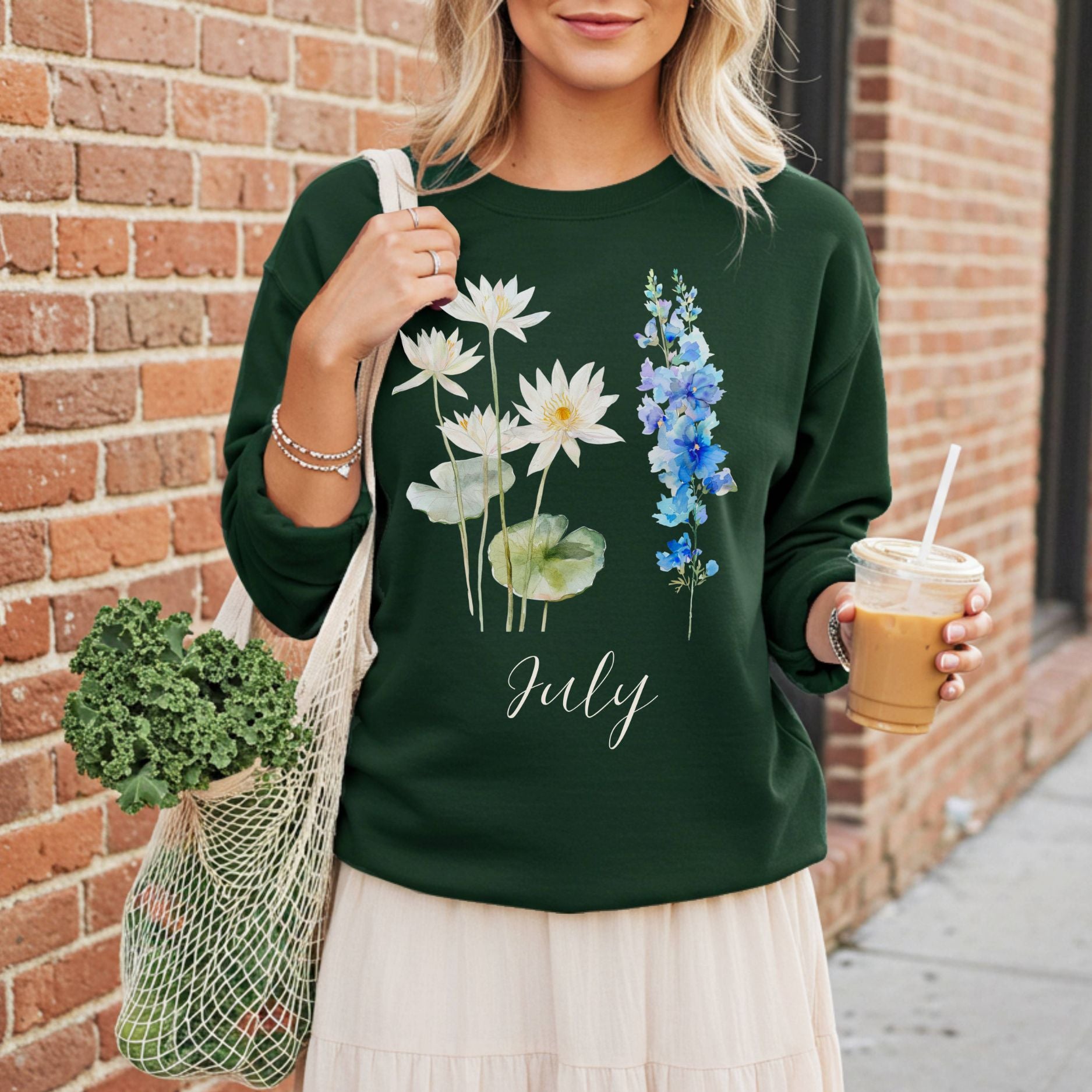 Woman in a dark green sweatshirt with watercolor waterlily and larkspur flowers, "July" script, holding coffee and a mesh bag.