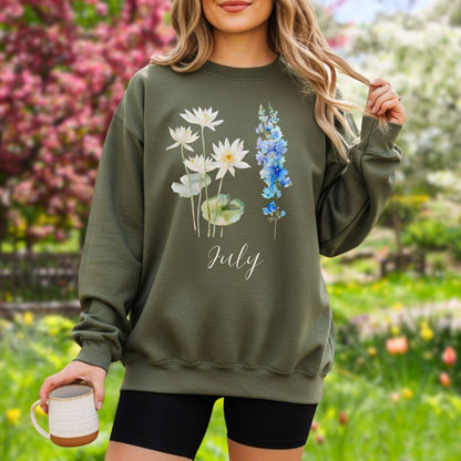 Woman in an olive green crewneck sweatshirt with watercolor waterlilies, larkspur, and "July" text.