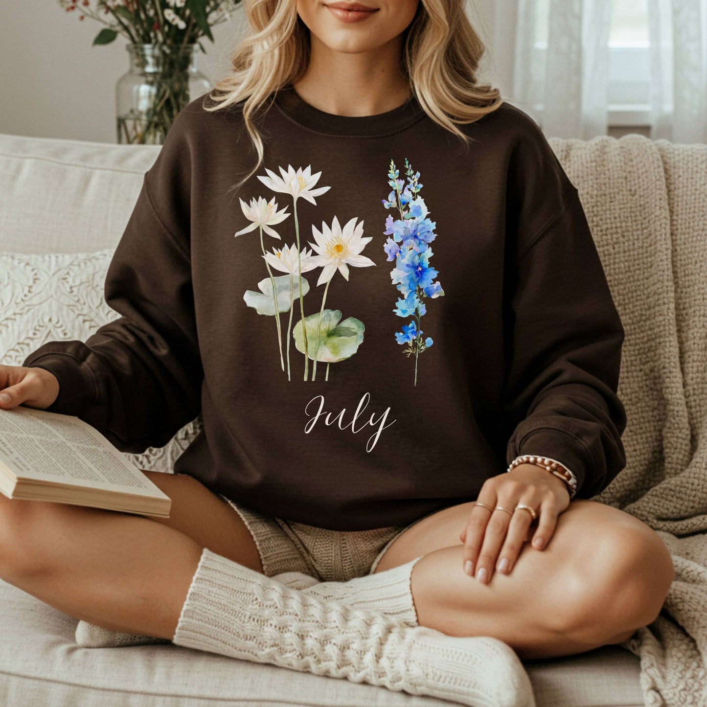 Woman in a dark brown crewneck sweatshirt with watercolor waterlily and blue larkspur flowers, plus "July" text.
