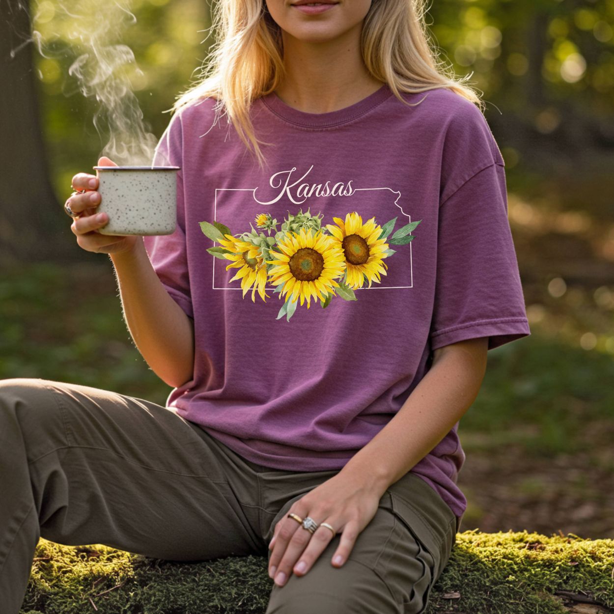 Garment-dyed plum t-shirt with Kansas state outline and vibrant yellow sunflowers, worn by a woman holding a mug.