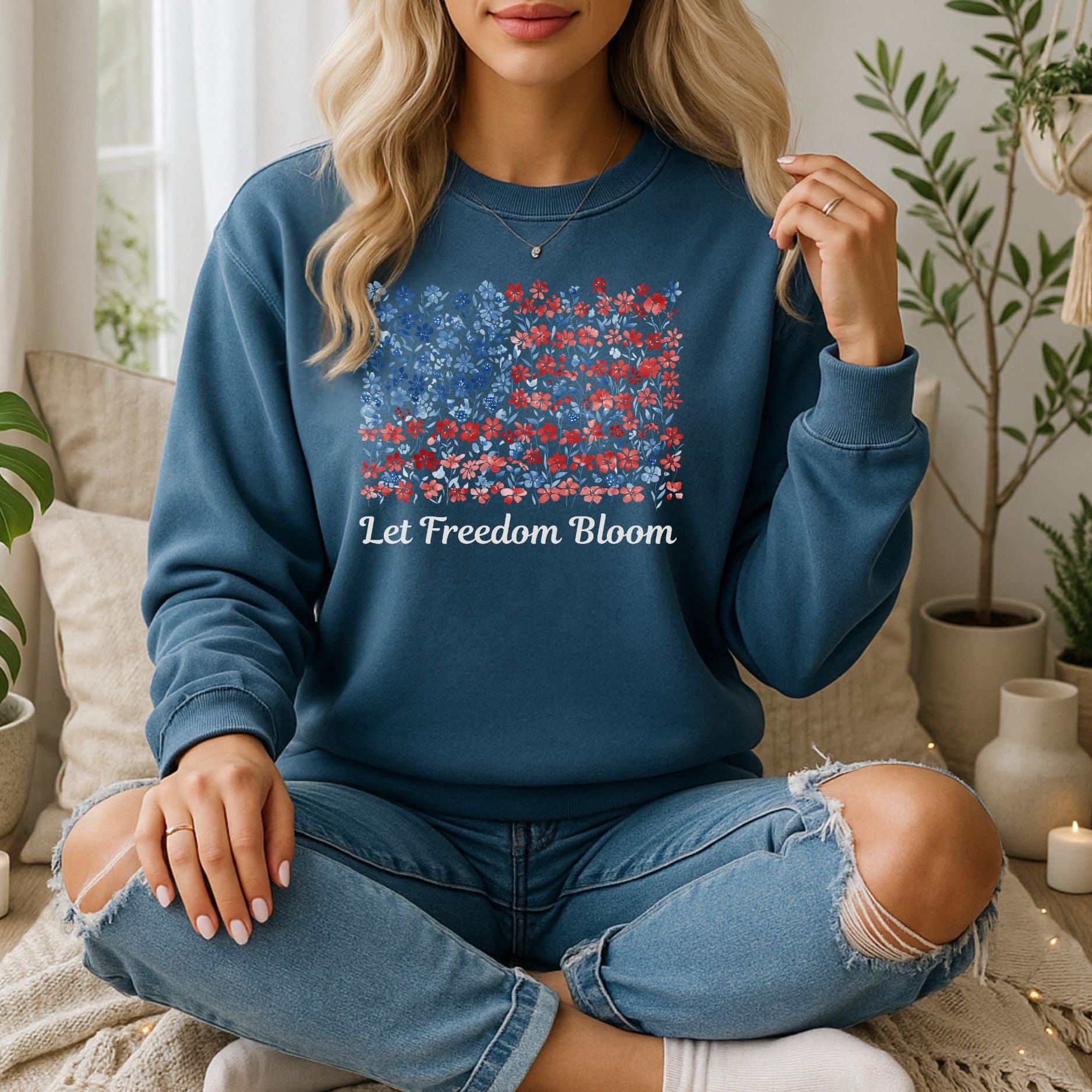 Blonde woman wearing a blue "Let Freedom Bloom" floral flag sweatshirt and ripped jeans, sitting indoors.