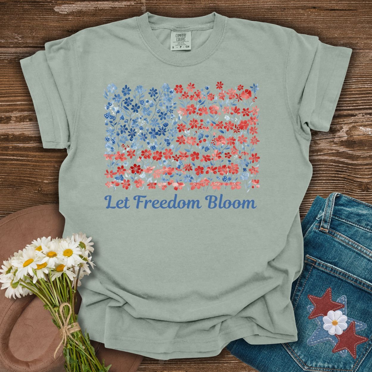 Bay-colored t-shirt with a floral USA flag design and "Let Freedom Bloom" text, on a wooden background.