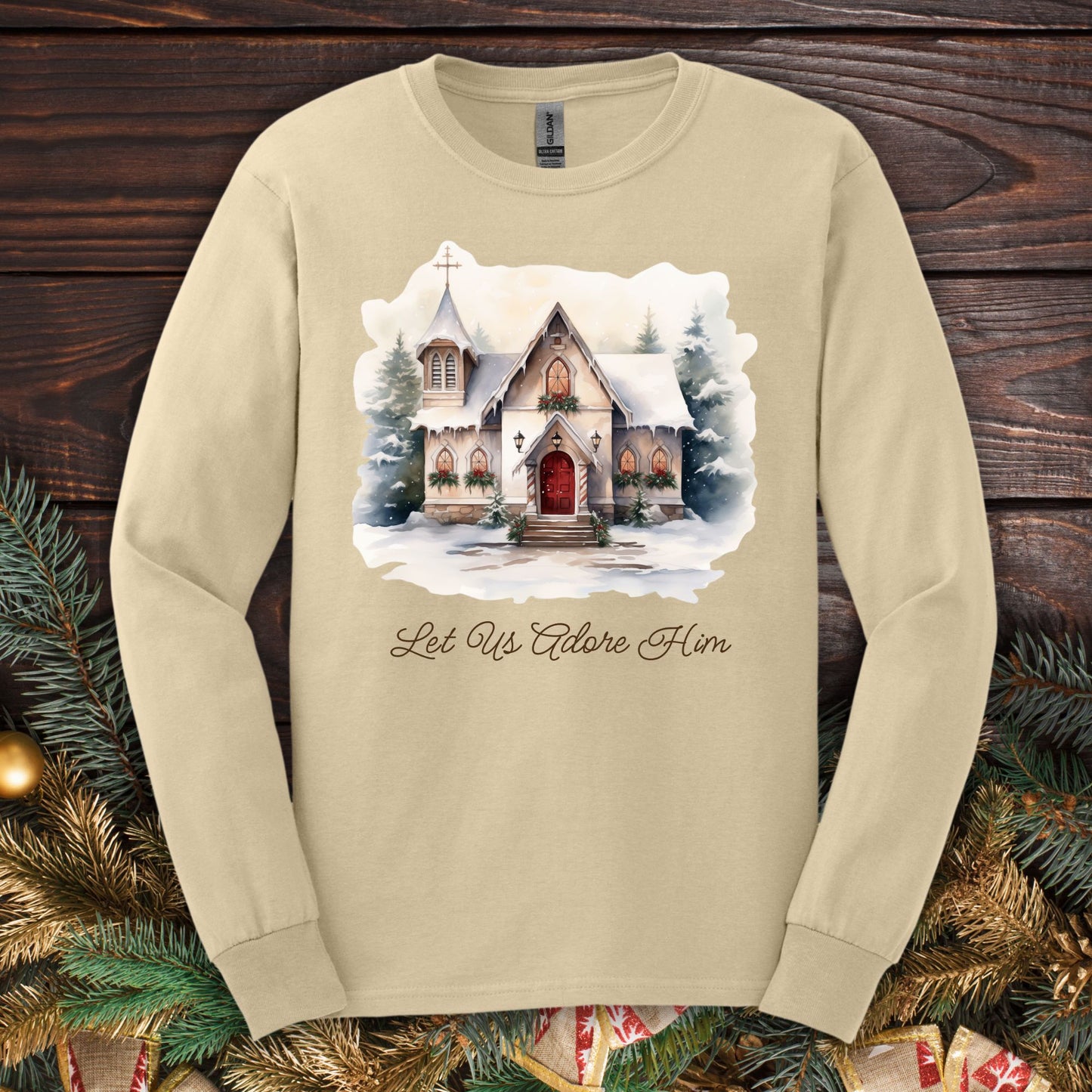 Sand long sleeve tee with a watercolor snow-covered chapel and "Let Us Adore Him" text.