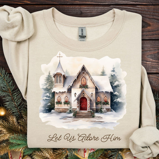 Sand Gildan sweatshirt with a snowy country chapel design, "Let Us Adore Him" text, and evergreen garland.