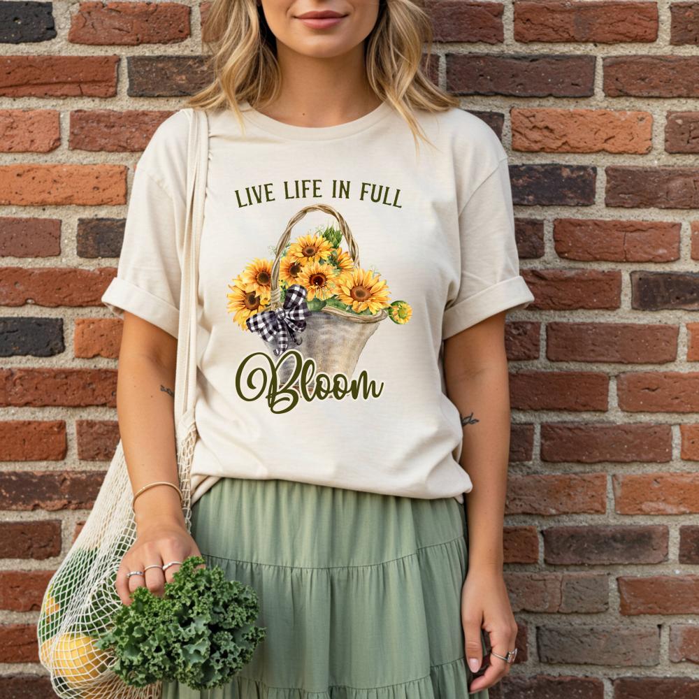 young woman wearing a t-shirt with a sunflower design and text that reads "Live Life in Full Bloom", standing against a brick wall.