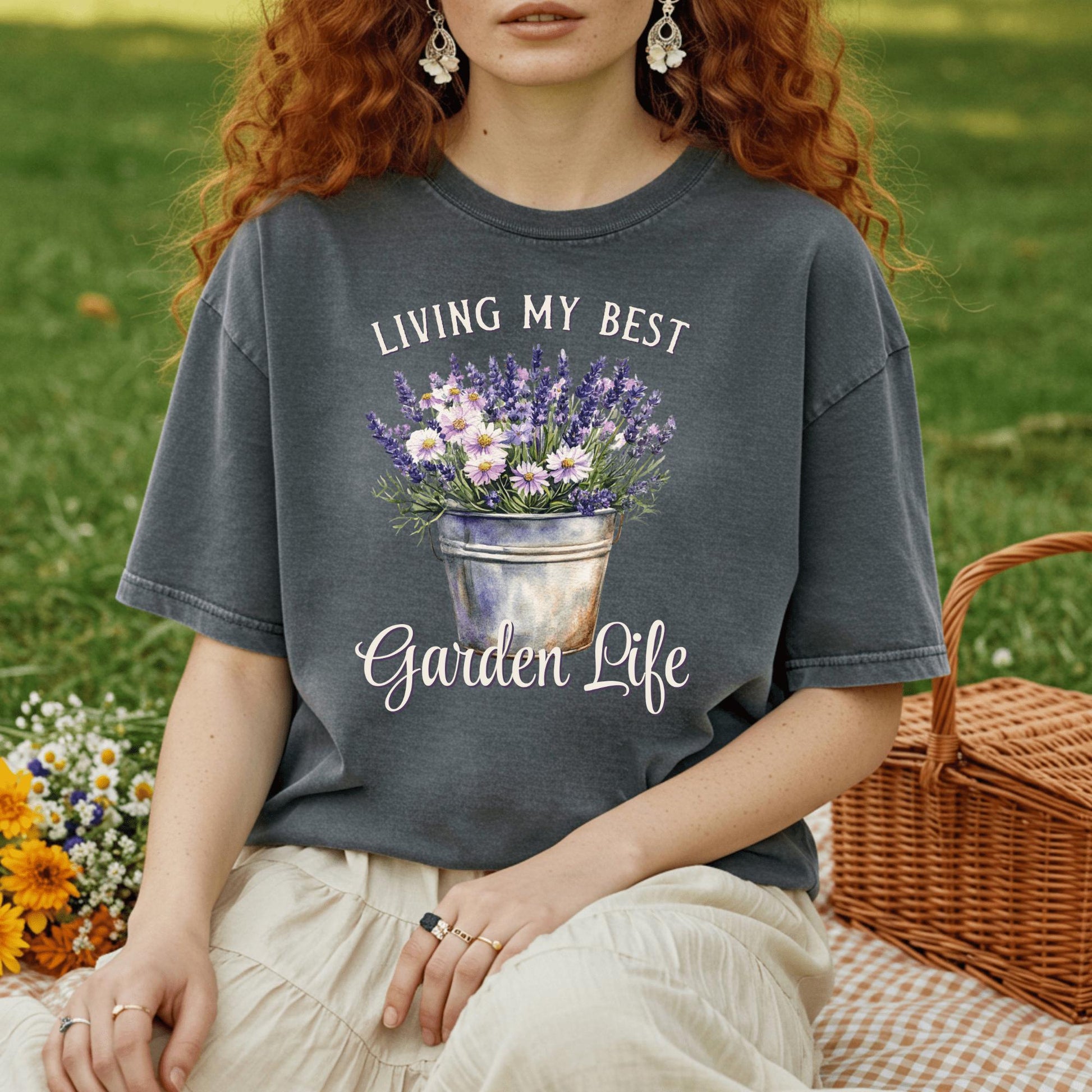 Woman in a charcoal gray "Living My Best Garden Life" t-shirt with a flower bucket graphic, sitting on a picnic blanket.