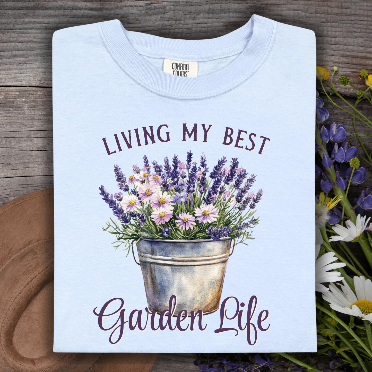 Folded hydrangea blue t-shirt with "Living My Best Garden Life" text and a lavender and daisy flower bucket graphic.