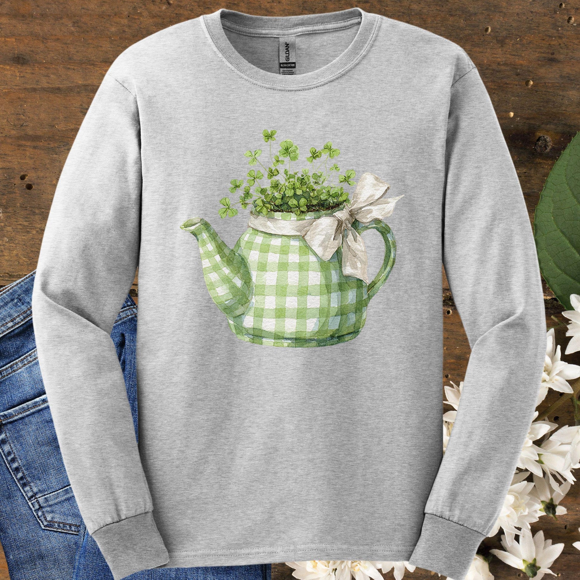 Ash long-sleeve tee with a green checkered teapot planter overflowing with clovers and a cream bow.